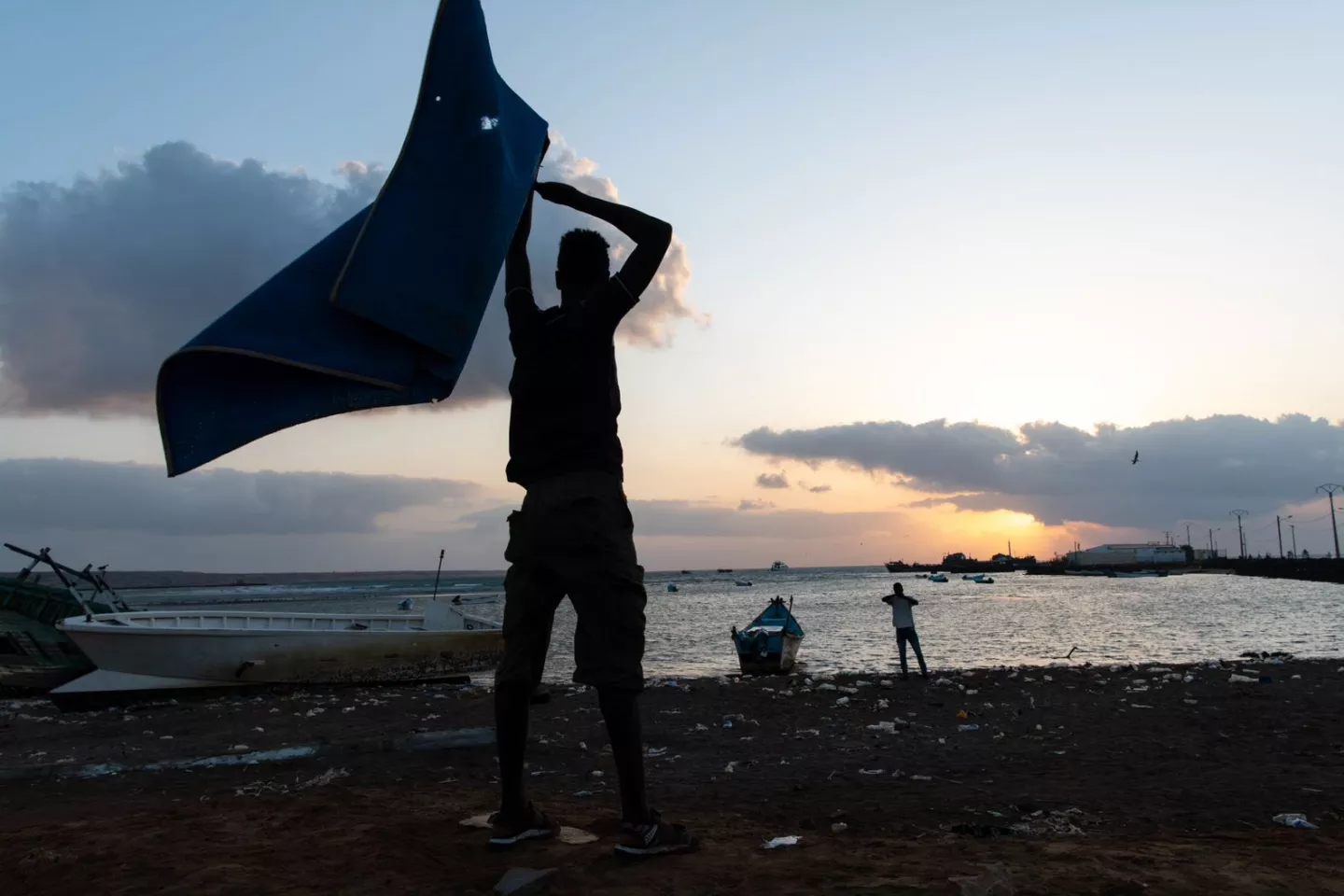 The silhouette of a man lifting a towel over his head, seen against the sunrise, near a lake.